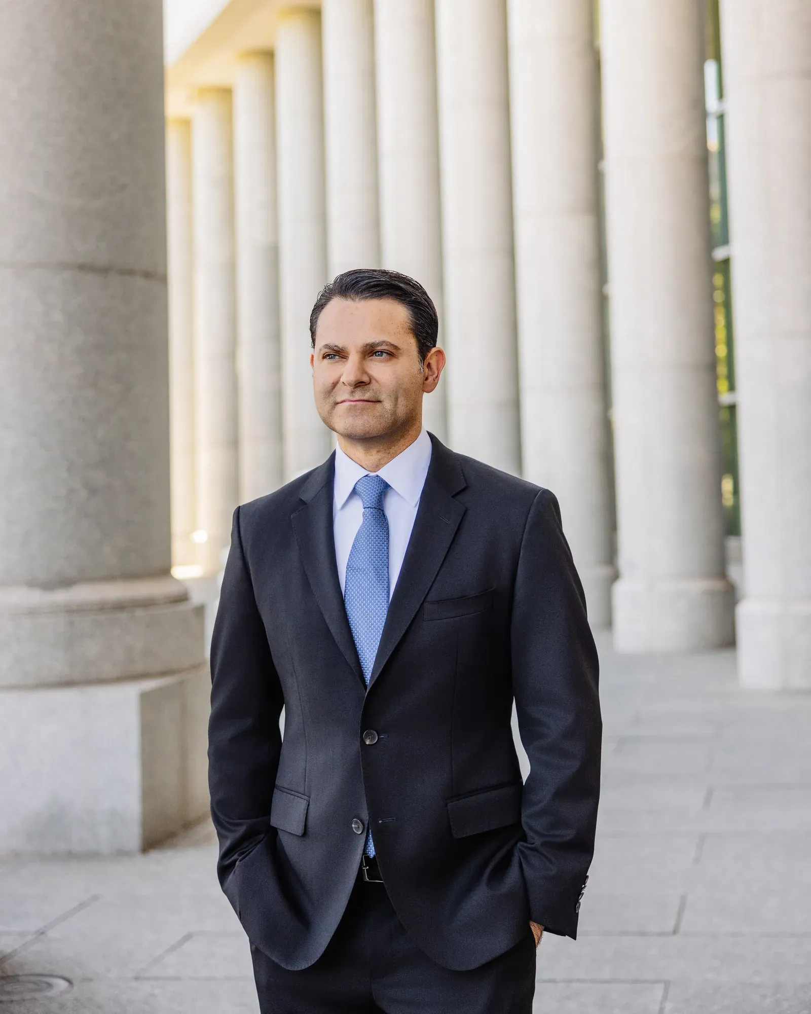 Mark Blechman in a charcoal suit and light blue tie, hands in his pockets, looking off to the side beneath a curving granite colonnade
